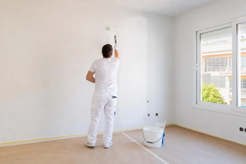 Pintor de espaldas pasando el rodillo en una pared de un salón vacío con el suelo protegido